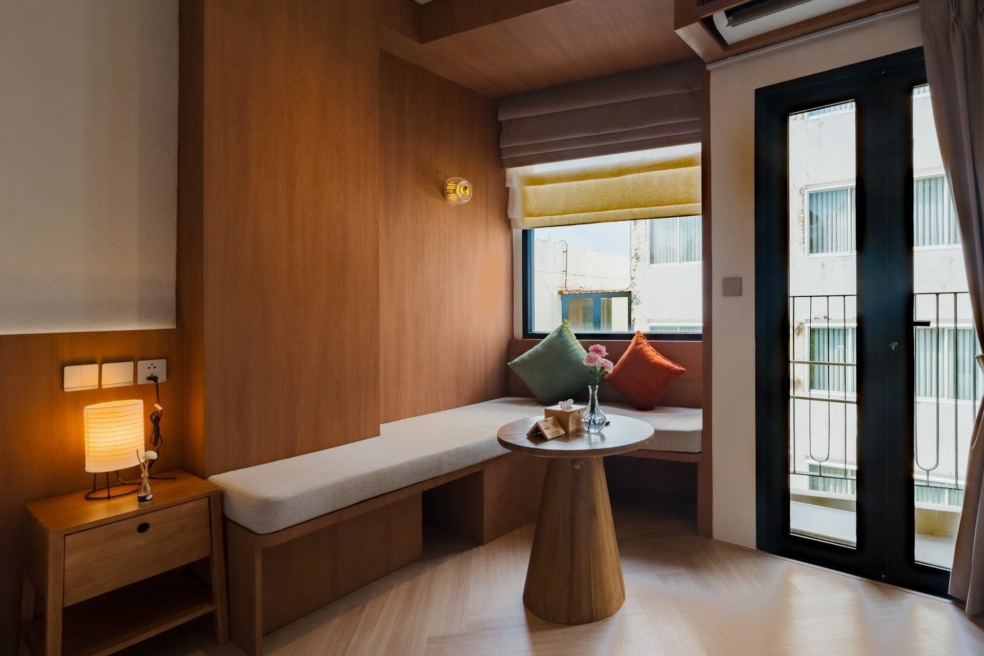 A hotel room with a wooden bench, a bedside table with a lamp, a round table with a vase, and a window with a view of buildings.