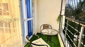 Ein Balkon mit künstlichem Grünbelag, zwei Stühlen, einem kleinen Tisch und einem Fenster mit blauen Vorhängen.