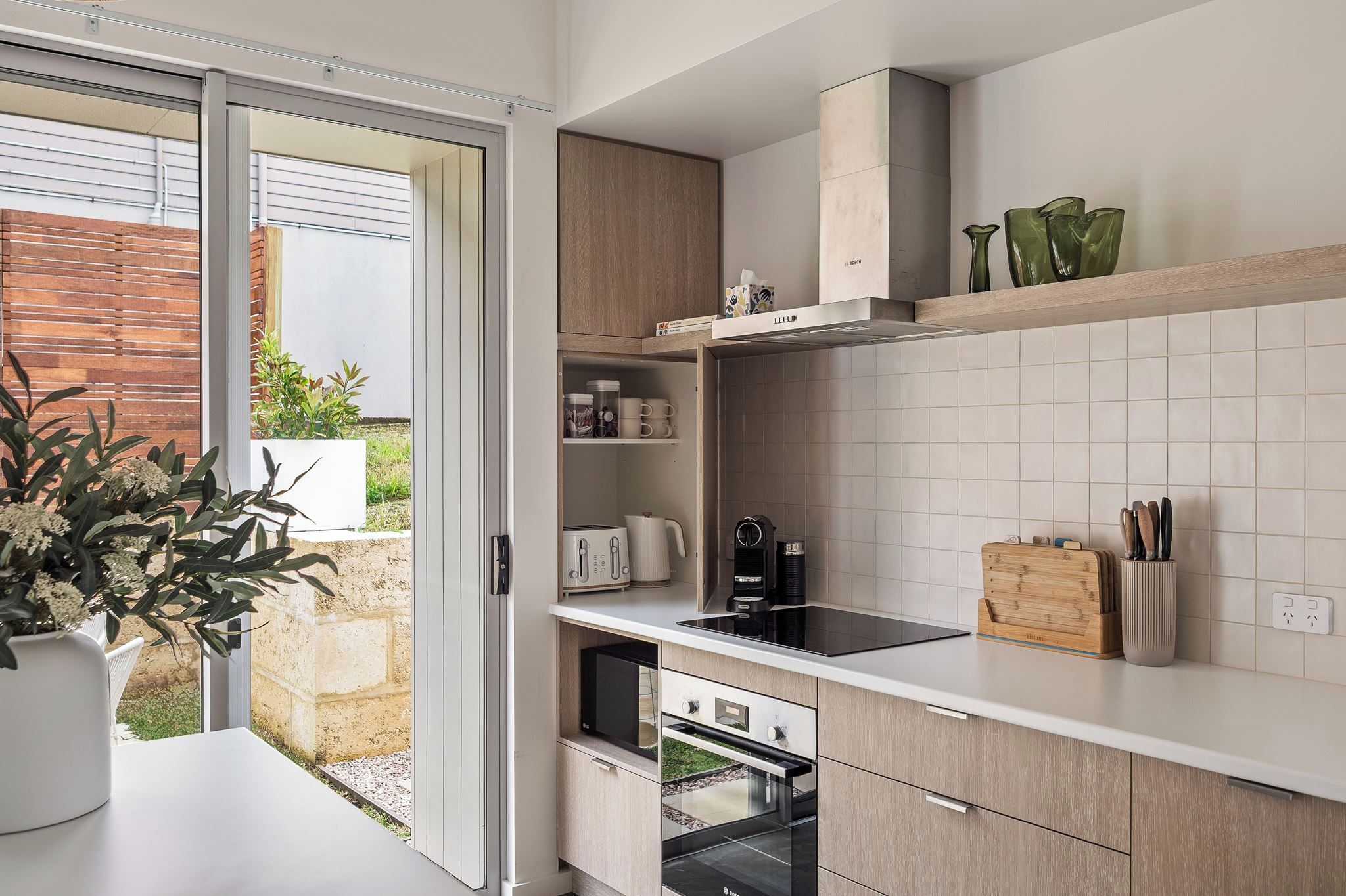 Une cuisine moderne dotée d’une porte-fenêtre coulissante donnant sur un patio, d’appareils électroménagers intégrés et d’un plan de travail en bois.