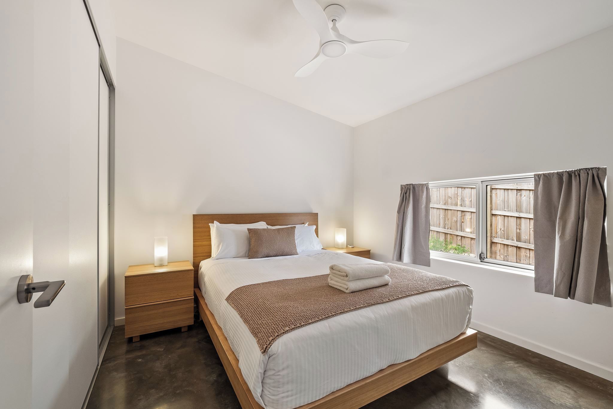 Une chambre à coucher comprenant un lit, des tables de chevet, un ventilateur de plafond, une fenêtre avec des rideaux et une vue sur l’extérieur.
