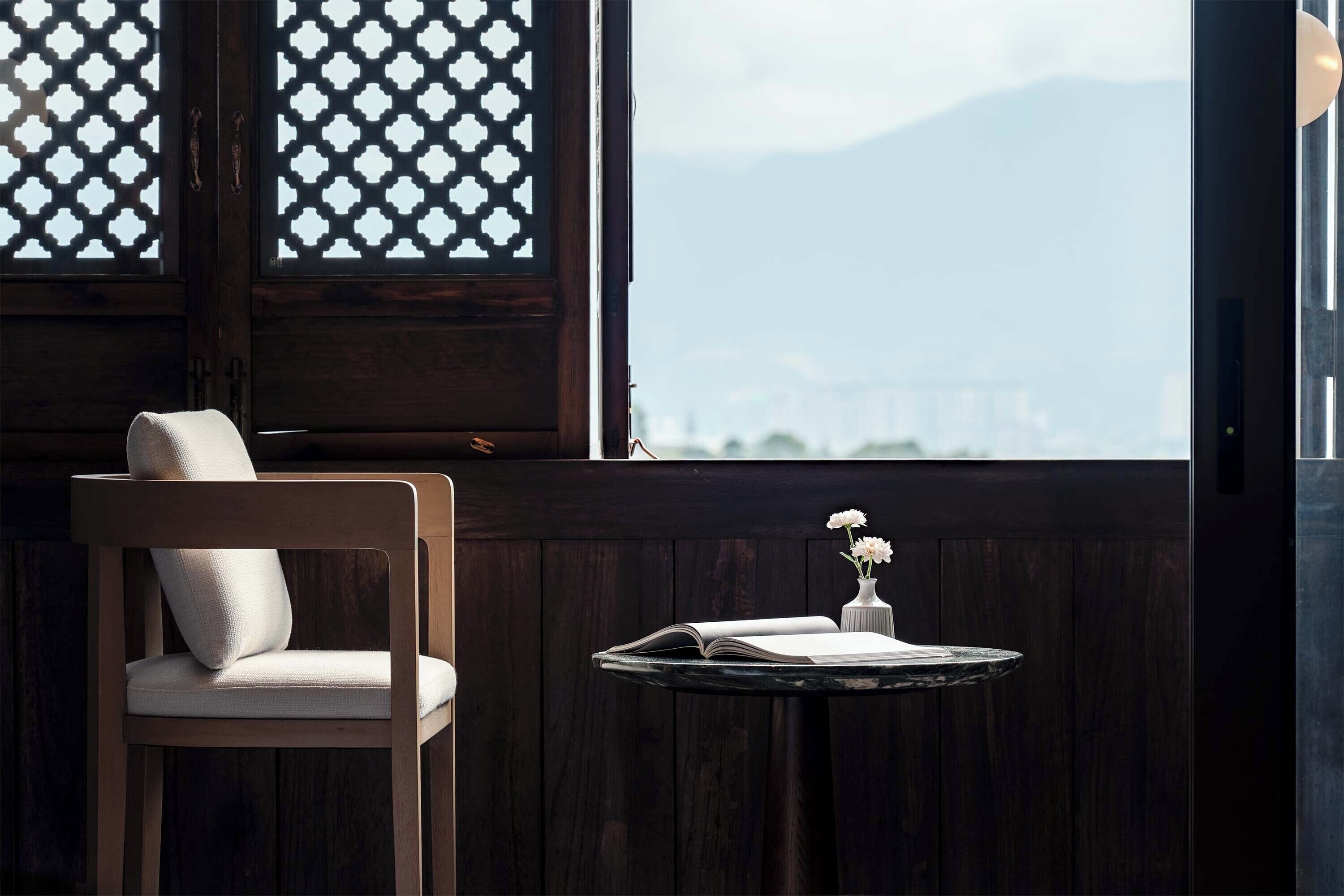 A wooden chair with a cushion, a small round table with a book and a vase of flowers, and a window with a lattice design.