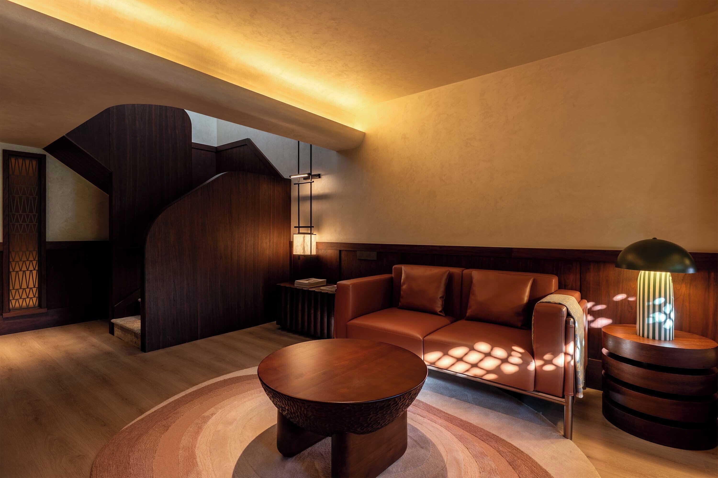A modern living room with a wooden staircase, a round coffee table, a leather sofa, and a side table with a lamp.