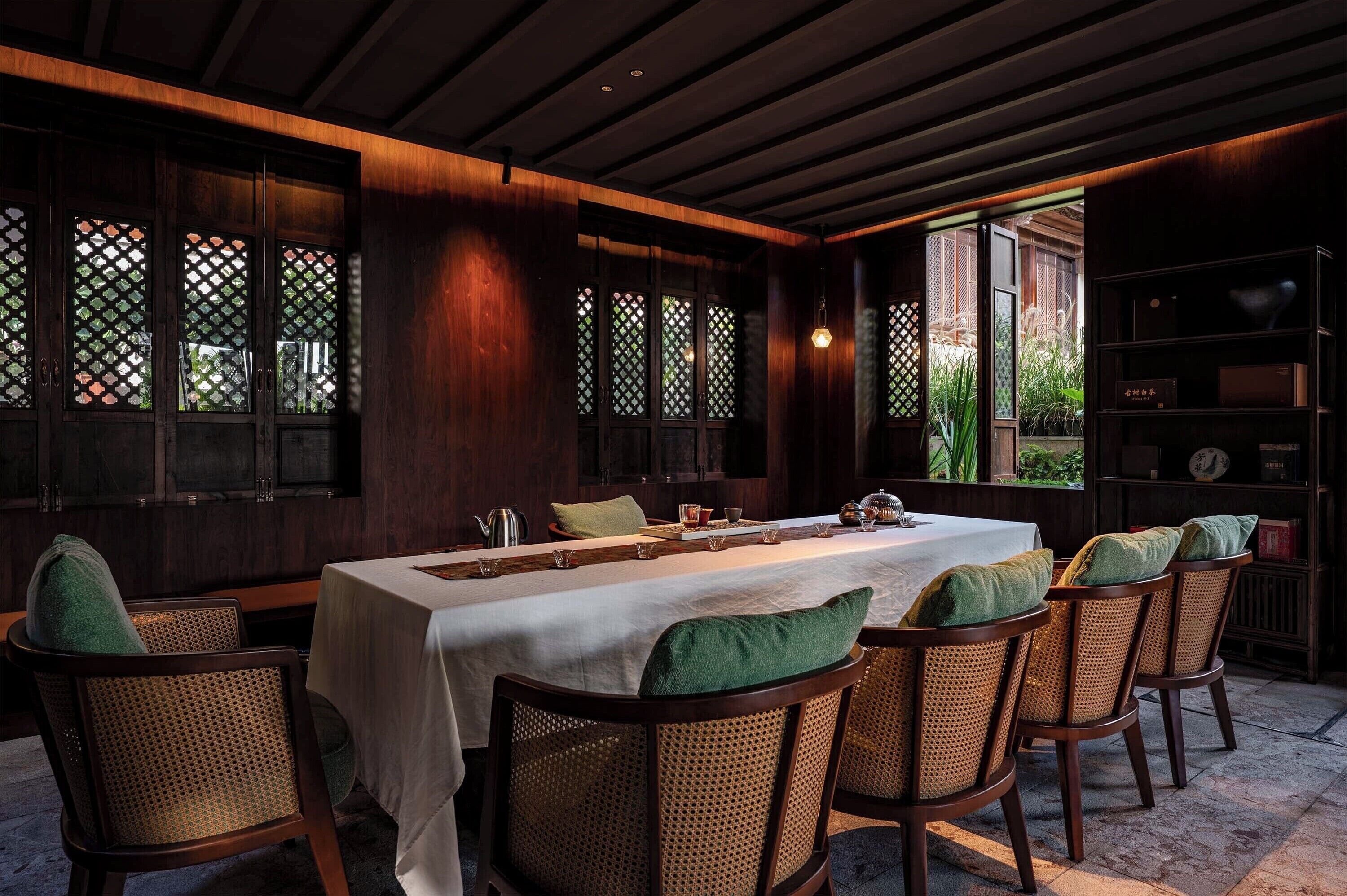 A dining area with a long table set for six, surrounded by wicker chairs with green cushions.