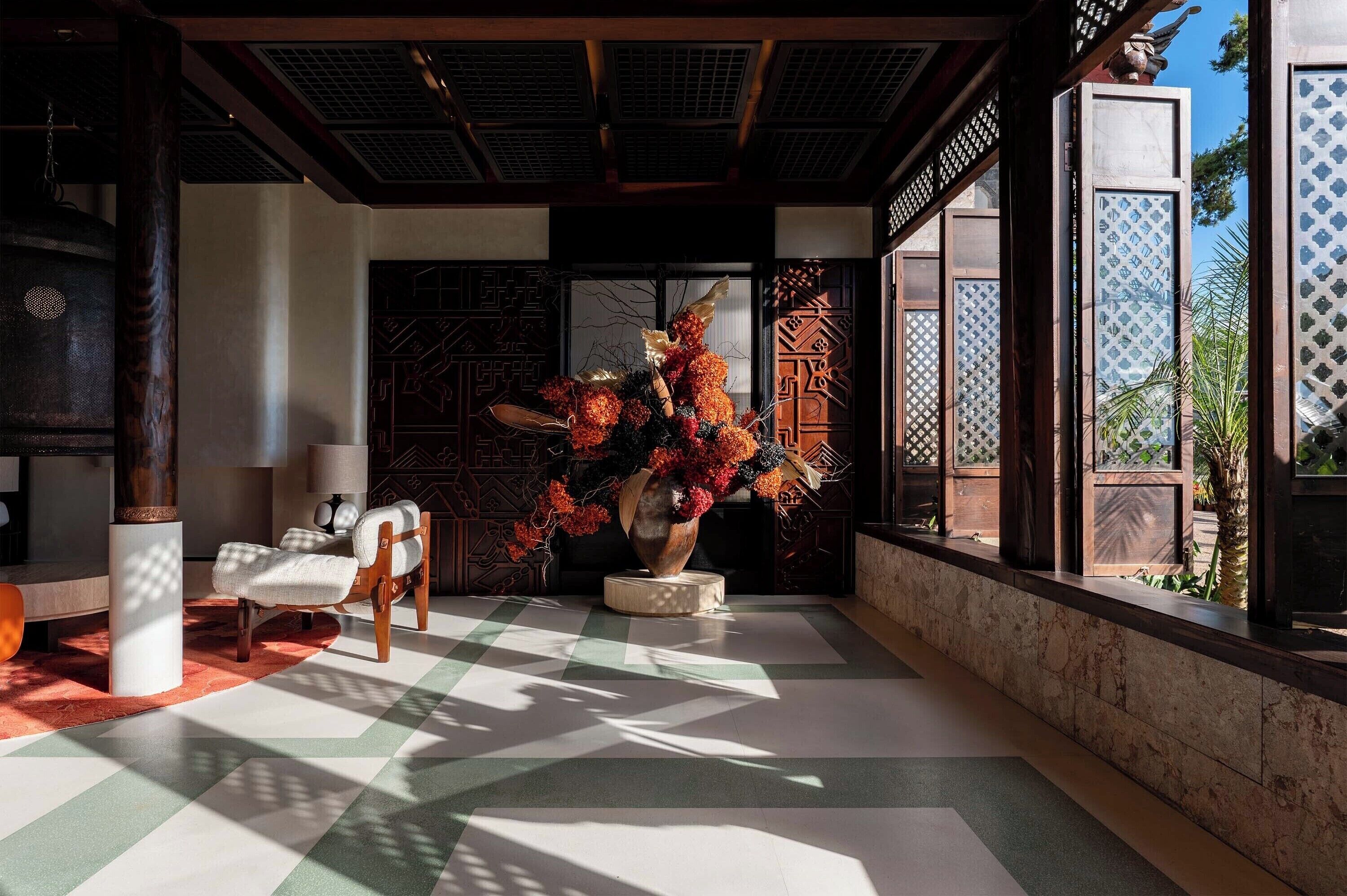 A traditional interior with a checkered floor, a wooden chair, a large vase with flowers, and a decorative wall.