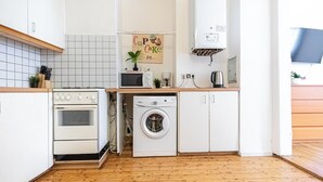 Een compacte keuken met een fornuis, magnetron en wasmachine, een kleine plant en een bord met de tekst 'Cupcake' aan de muur.