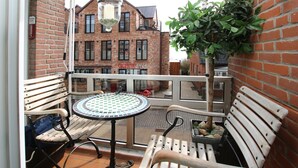 Un balcon avec une table et deux bancs en bois, une plante en pot et, au fond, un bâtiment en briques.