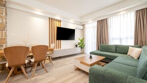 Een moderne woonkamer met een groene bank, een houten eettafel en stoelen. Een flatscreen televisie is aan de muur bevestigd, en er staat een plant op de bijzettafel.