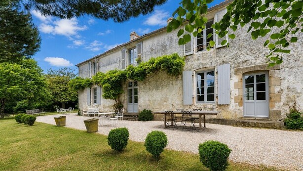 A stone building with a gravel driveway, a wooden table with chairs, and trimmed bushes.