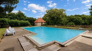 A rectangular swimming pool with a wooden deck and lounge chairs, surrounded by greenery and a stone building with a red roof.
