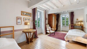 A room with wooden flooring, a bed, a chair, a table, a window with curtains, and framed pictures on the wall.