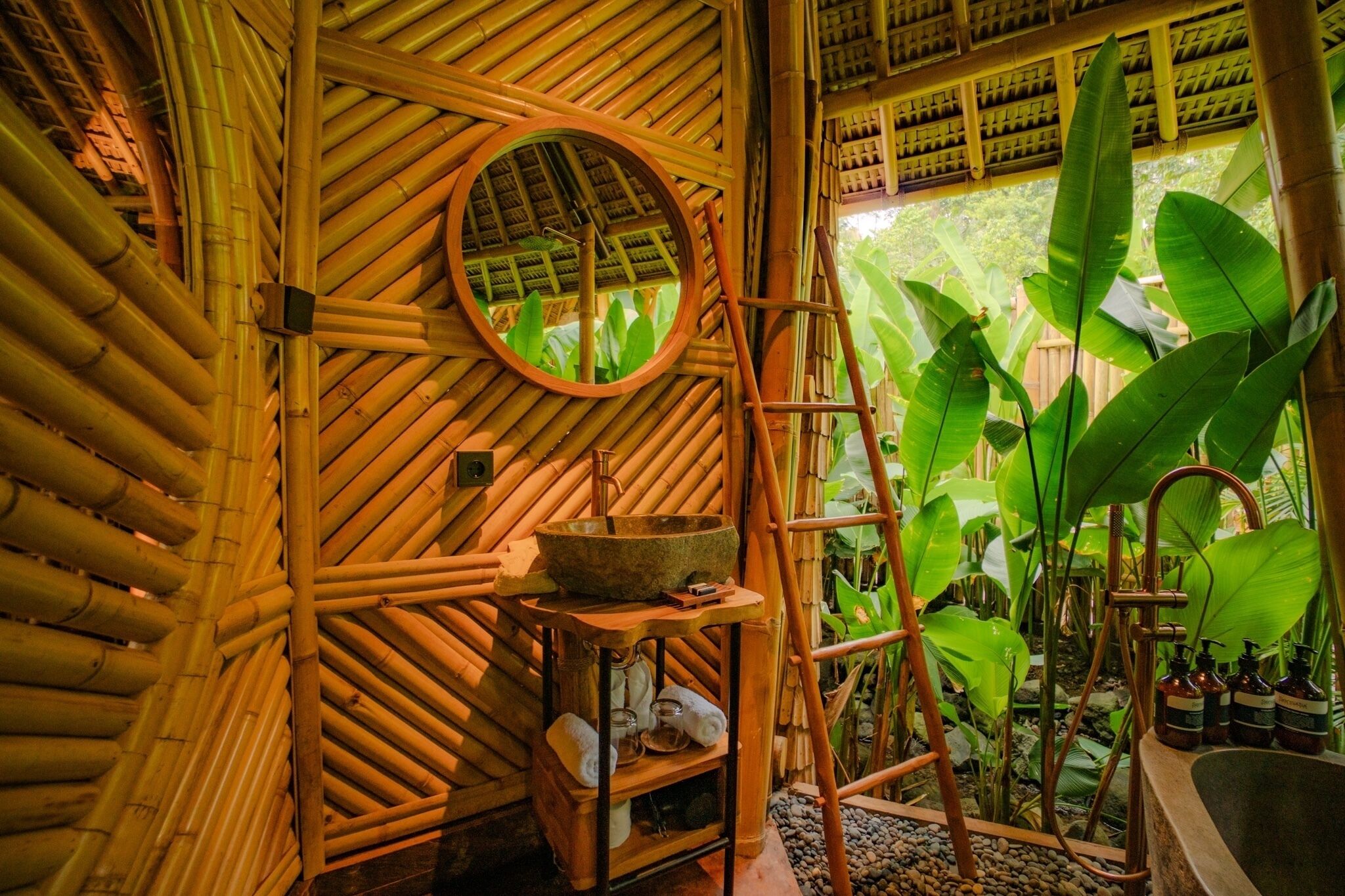 A bamboo room with a round mirror, a stone basin, a wooden shelf with towels, and a ladder.