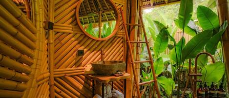 A bamboo room with a round mirror, a stone basin, a wooden shelf with towels, and a ladder.