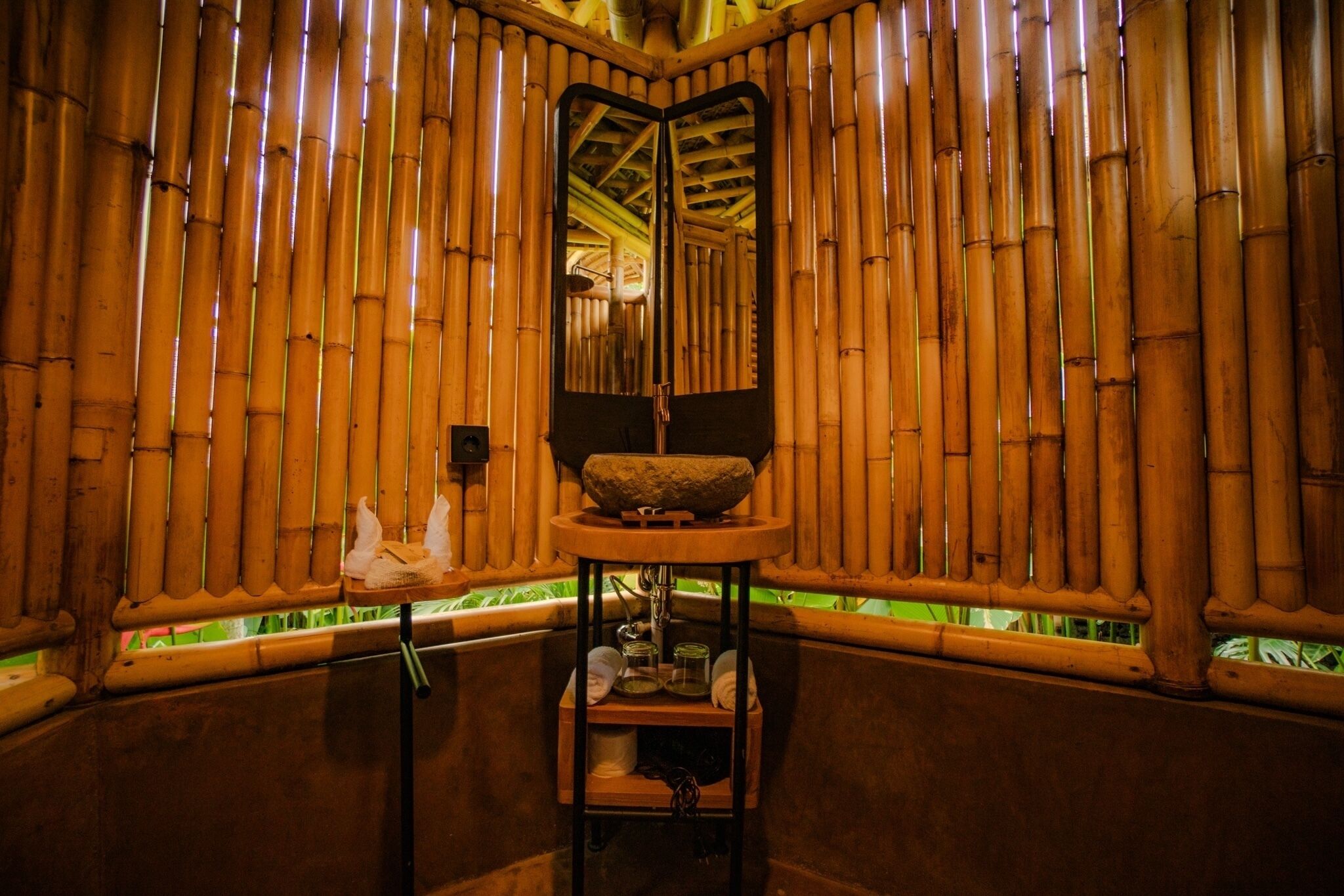 Ein Zimmer mit Bambuswänden, einer zentralen Steinarchitektur und einem kleinen Tisch mit Handtüchern und Pflegeprodukten.