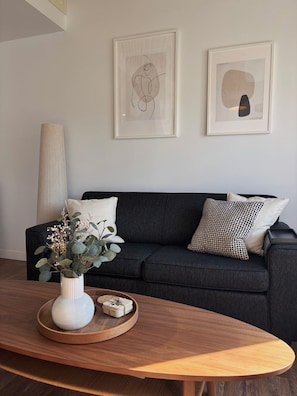 Uma sala de estar moderna com um sofá cinza escuro, um vaso branco com flores e duas obras de arte abstratas na parede.