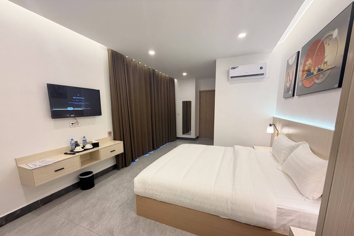 A modern hotel room with a large bed, a flat-screen TV mounted on the wall, a bedside table with a lamp, and an air conditioning unit.
