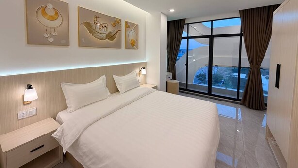 A modern hotel room with a large bed, a nightstand, a wall-mounted lamp, and artwork above the bed. - The Sky Plus (Sihanoukville)