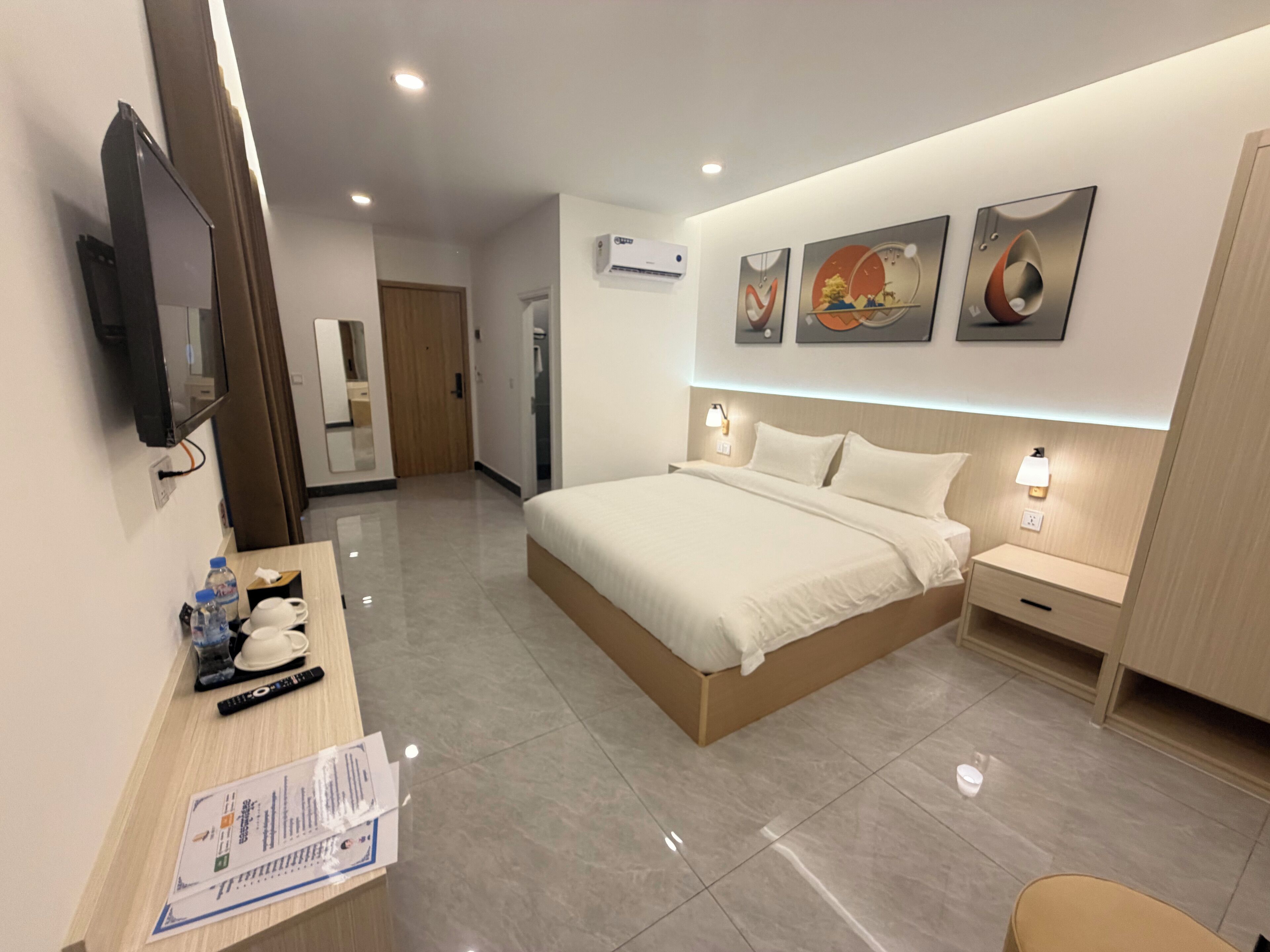 Una habitación de hotel moderna con una cama grande, mesitas de noche con lámparas, un televisor de pantalla plana en la pared y una obra de arte sobre la cama.