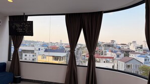 A room with a large window, curtains, a blue sofa, and a TV on the wall. - The Sky Plus (Sihanoukville)