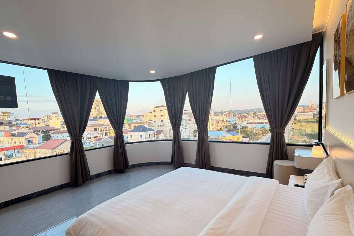 A hotel room with a large bed, a bedside table, a lamp, and a view of a cityscape through large windows with curtains.