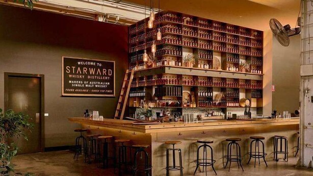 Zona de bar con un cartel que dice 'Welcome to Starward Whisky Distillery', estantes llenos de botellas y taburetes de bar.