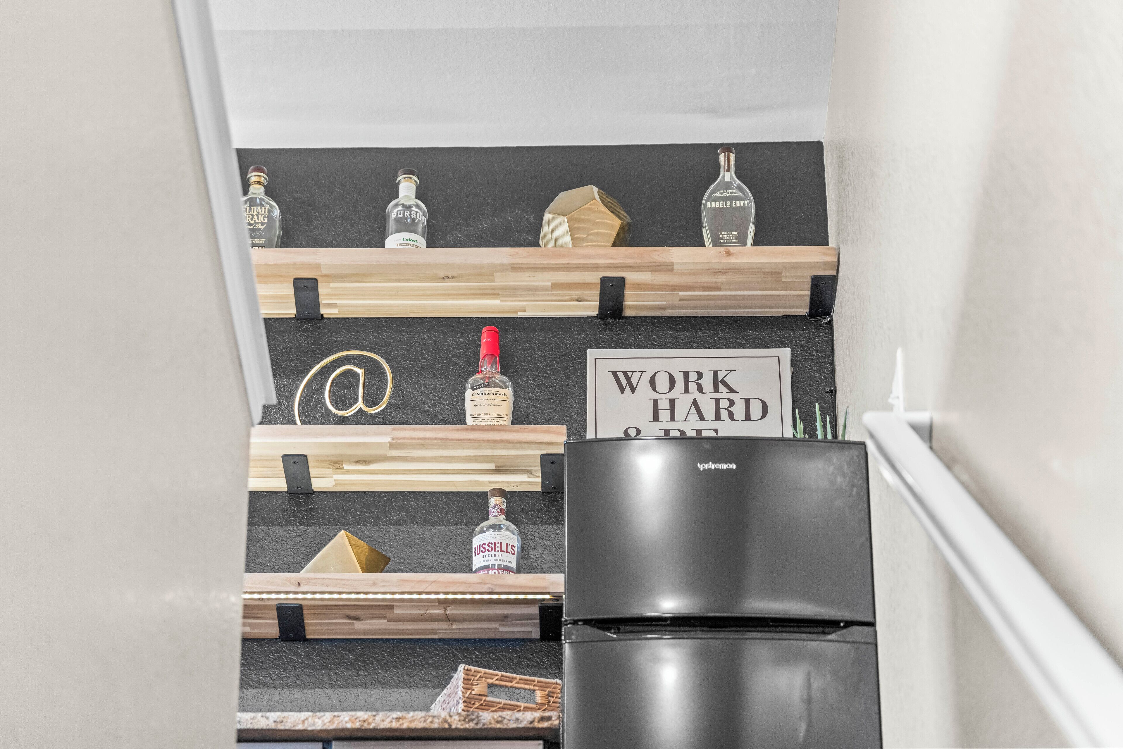 A kitchen with a black refrigerator, wooden shelves holding bottles, and a sign that reads "WORK HARD & BE KIND".