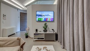 Un soggiorno moderno con una TV a schermo piatto montata a parete, un divano grigio, un tavolino da caffè con oggetti decorativi, una pianta e una porta in vetro che dà su un'altra stanza.