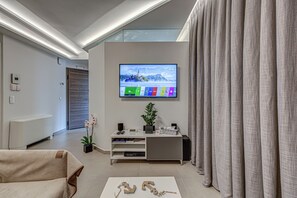 Ein modernes Wohnzimmer mit Flachbildfernseher an der Wand, einem grauen Sofa, einem Couchtisch mit Dekoration, einer Pflanze und einer Glastür zu einem anderen Raum.