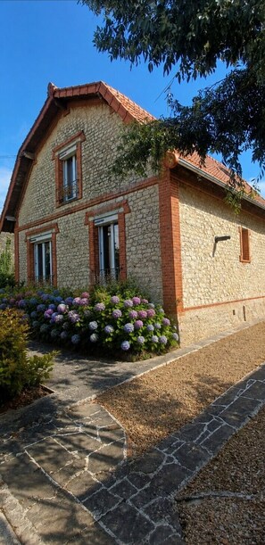 Een stenen huis met een pannendak, een stenen pad en een bloembed met hortensia’s.