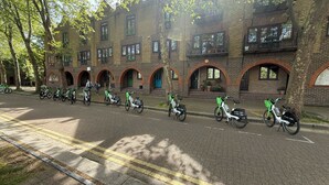 Un bâtiment en briques, doté de nombreuses fenêtres et d’entrées en arc de cercle, bordé d’arbres et où sont stationnés des scooters électriques.