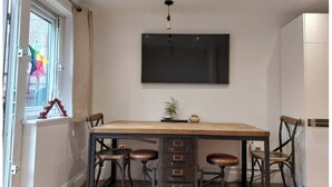 Un coin repas comprenant une grande table, quatre chaises, un téléviseur fixé au mur et une fenêtre avec des rideaux.