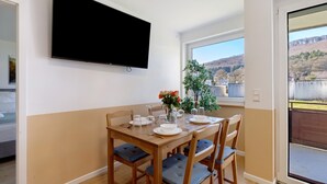 Uma área de refeições com uma mesa posta para quatro pessoas, uma TV de tela plana na parede e vista para uma encosta através de uma janela.