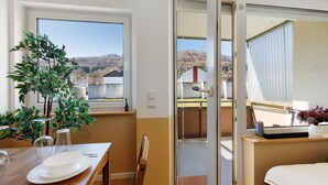 Uma área de refeições com uma mesa posta para dois, uma planta e vista do exterior através de uma porta de vidro deslizante.
