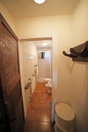A narrow hallway with a wooden door, a white wall, a tiled floor, a white trash bin, and a bathroom visible in the background.