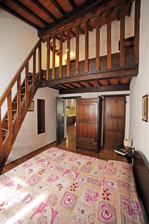A bedroom with a wooden staircase, a large bed with a patterned bedspread, and a view of the kitchen through an open door.