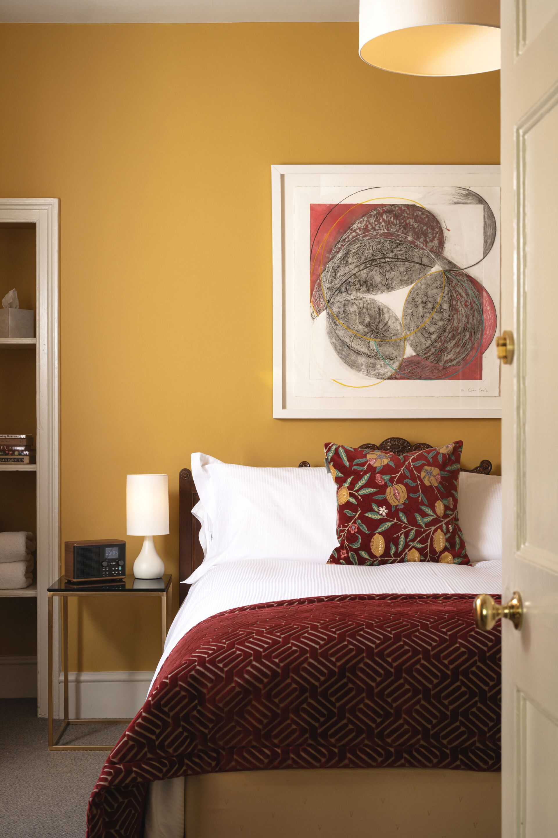 A hotel room with a bed, a nightstand with a lamp, a framed abstract painting, and a door.