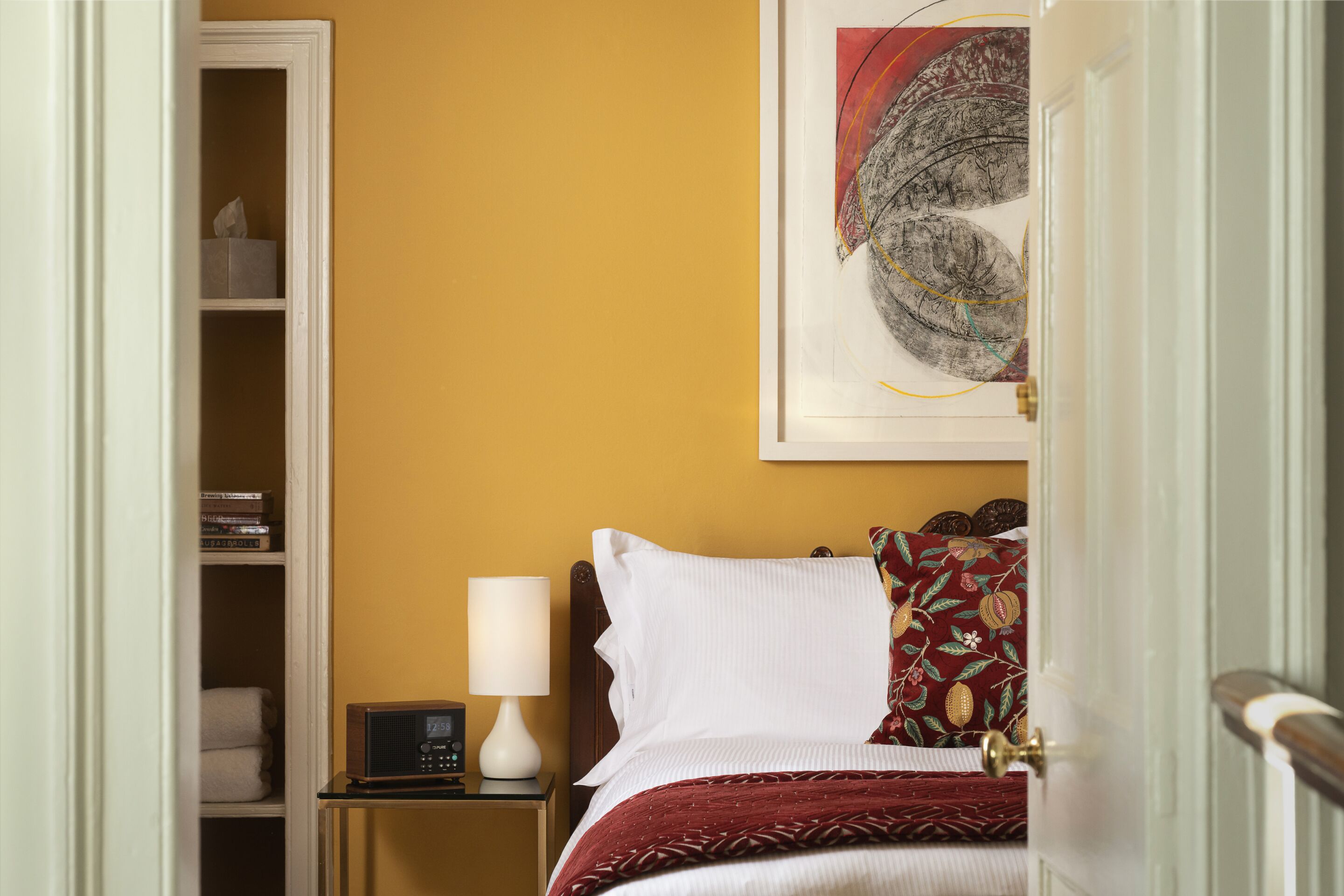 Habitación de hotel con cama, mesita de noche con lámpara, estantería y un cuadro en la pared.