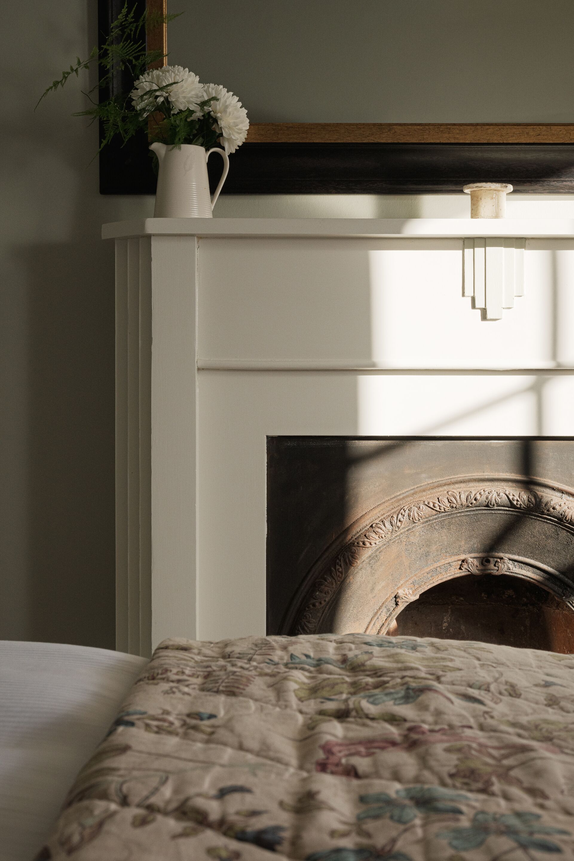 A bedroom with a white bed, a floral quilt, a white pitcher with flowers, and a fireplace.