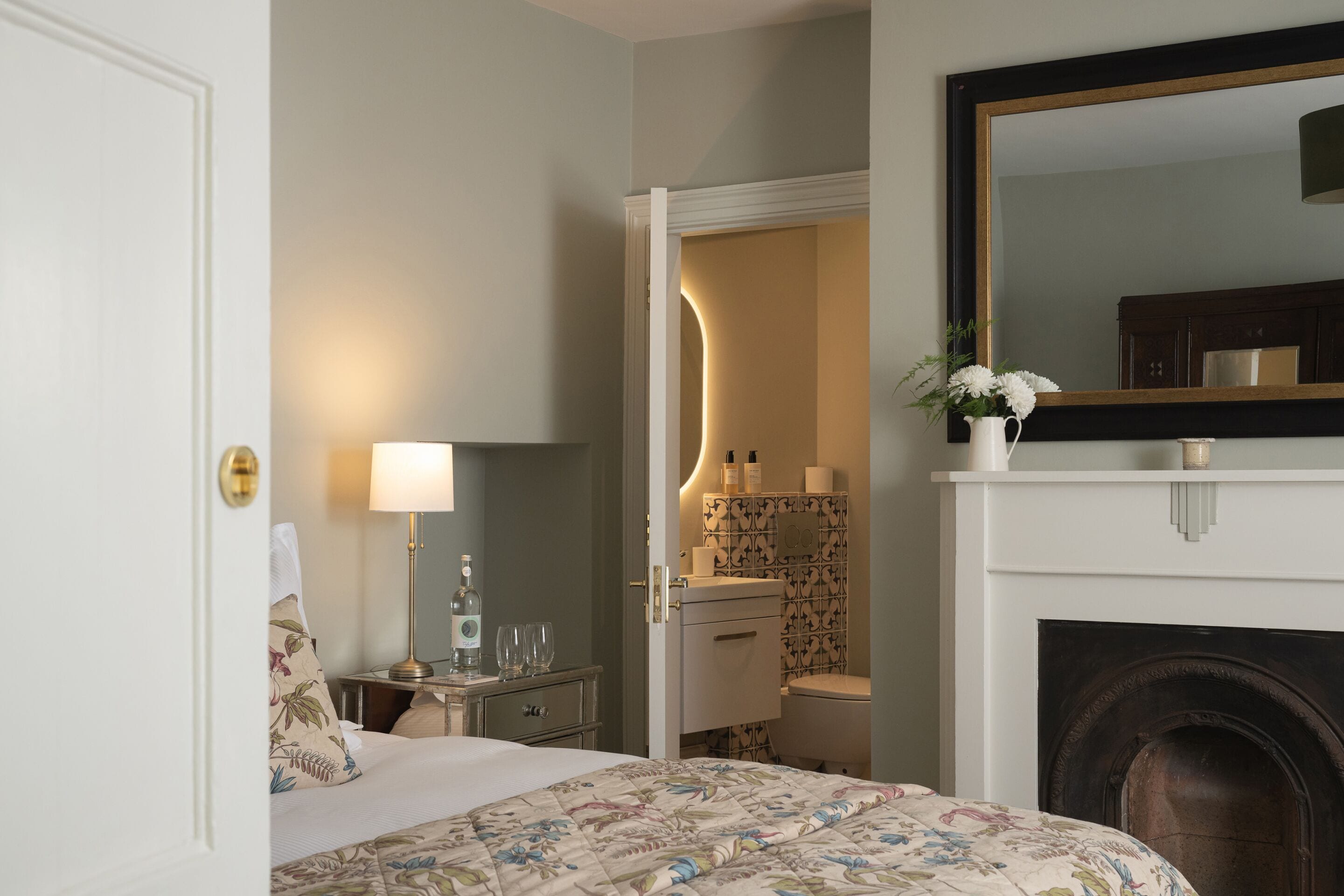 A hotel room with a bed, bedside table, lamp, and a fireplace.