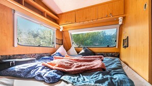Un dormitorio acogedor en una caravana, con cama, almohadas, manta y ventana con vistas a la vegetación.