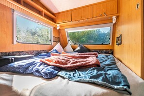 Quarto aconchegante de um motorhome, com cama, travesseiros, cobertor e janela com vista para o verde.