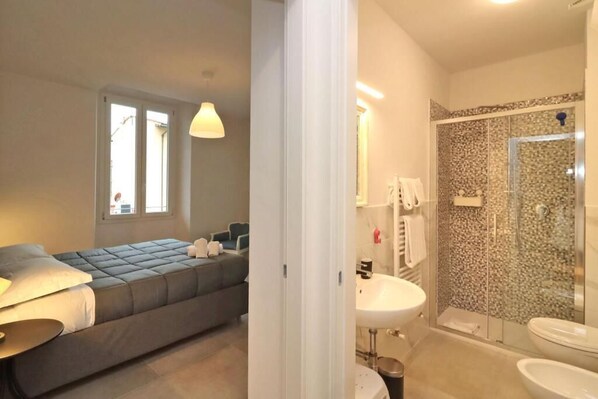 A modern bedroom with a sofa bed, a bedside table, and a window. Adjacent is a bathroom with a shower, sink, and toilet.