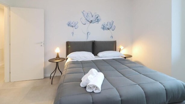 A neatly made bed with a grey quilt, two white pillows, and a rolled white towel on the bed. There are two bedside tables with lamps, and a wall with a floral painting. - Rose Suite in Florence (Florence)