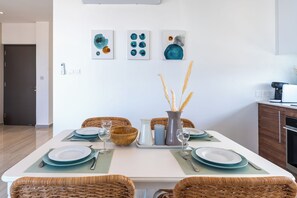A dining area with a white table set for four, featuring teal plates and silverware, a vase with dried plants, and abstract art on the wall. - Thálassa 2-BR Apt. & Balcony in Larnaca (Larnaca)