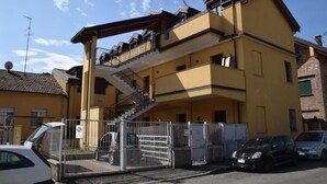 Un immeuble jaune à plusieurs étages, doté d’un escalier central, d’une entrée fermée par un portail et d’un espace de stationnement où sont garées des voitures.