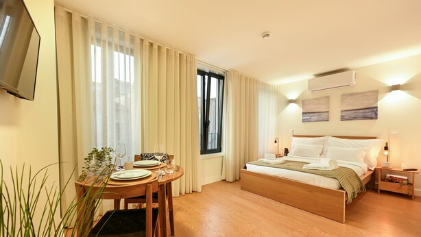 A hotel room with a bed, bedside table, lamp, curtains, a TV mounted on the wall, and a window. - ON Apartments - MA62 (Porto)