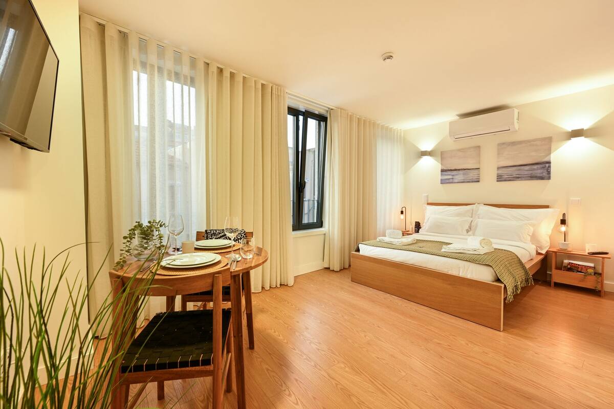 A hotel room with a bed, bedside table, lamp, curtains, a TV mounted on the wall, and a window.