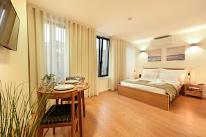 Quarto de hotel com cama, mesa de cabeceira, abajur, cortinas, televisão fixada na parede e uma janela.