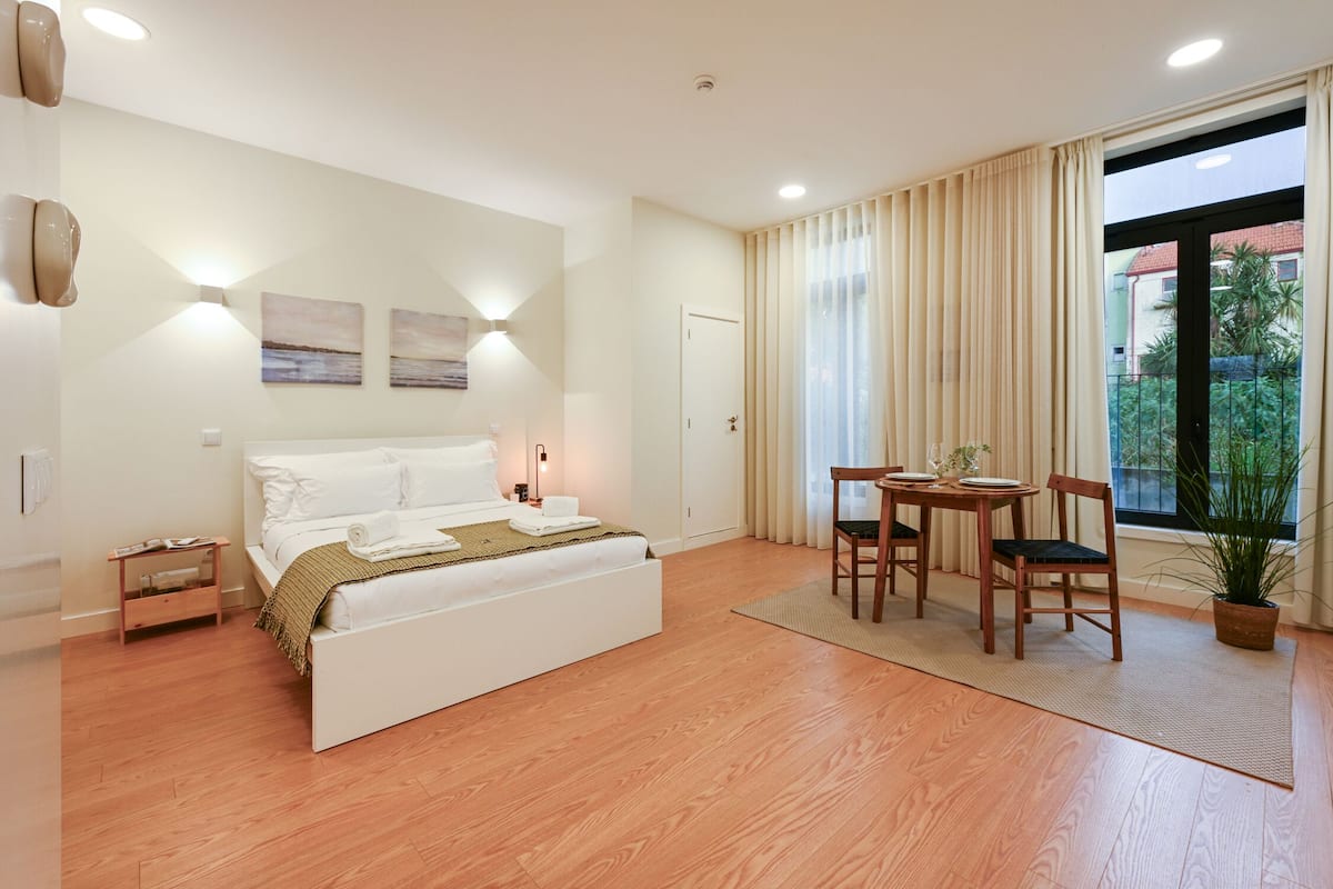 A bedroom with a bed, bedside table, lamp, and a view of the outside.