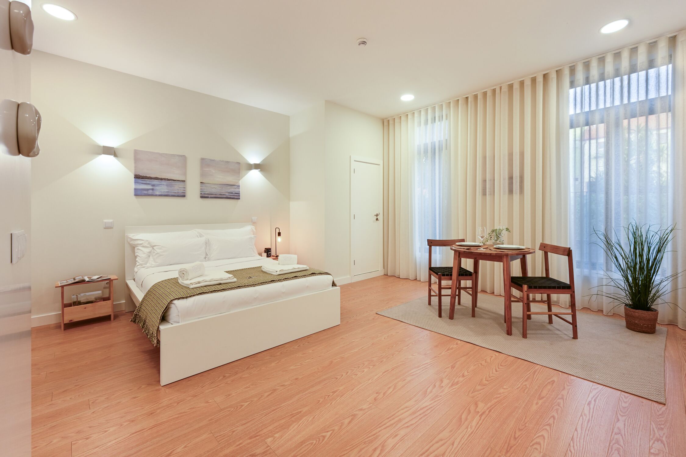 A bedroom with a bed, bedside table, lamp, curtains, and a potted plant.