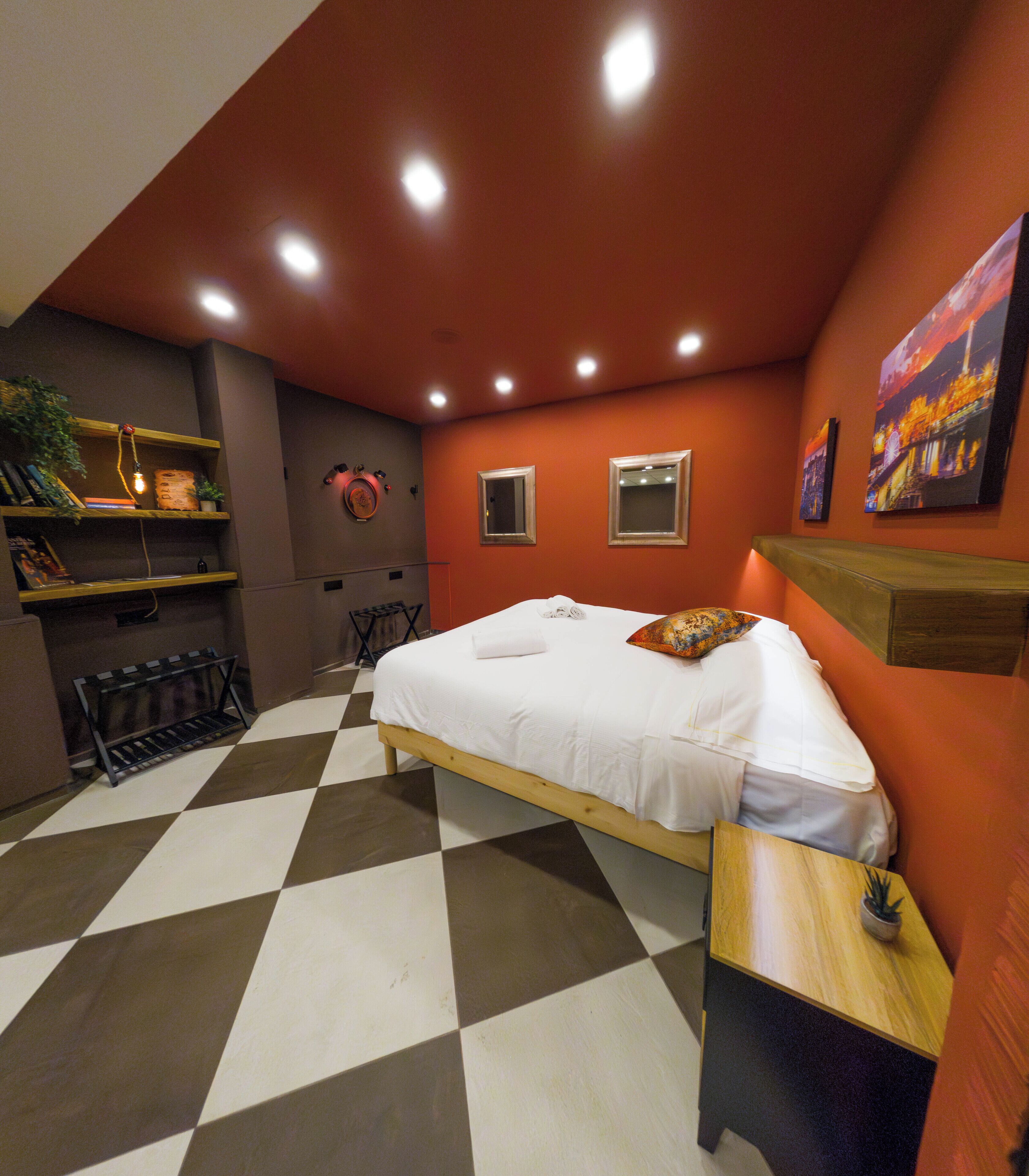 Quarto com chão xadrez, cama com roupa de cama branca, duas mesas de cabeceira, uma pequena estante com livros, uma planta em vaso e um quadro na parede.